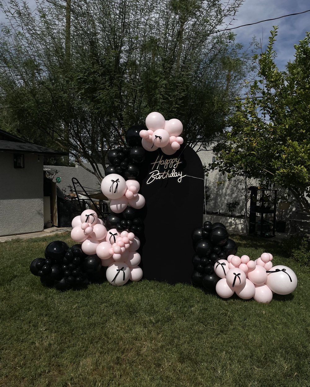 A tall black arched backdrop with a neon Happy Birthday sign and pink and black balloons tied with black ribbon bows