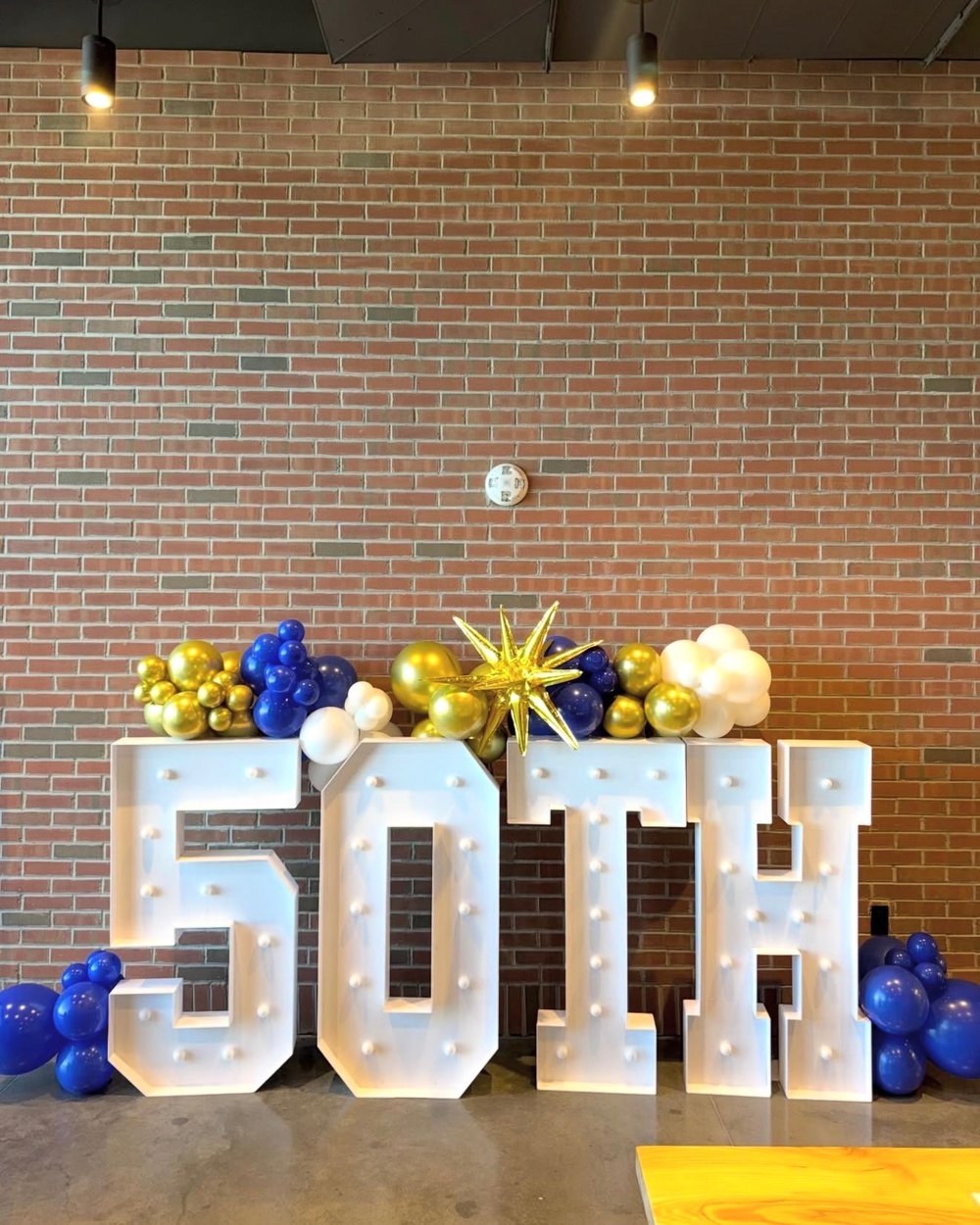 Light-up 50TH letters with navy, gold and white balloons and a gold starburst against a brick wall
