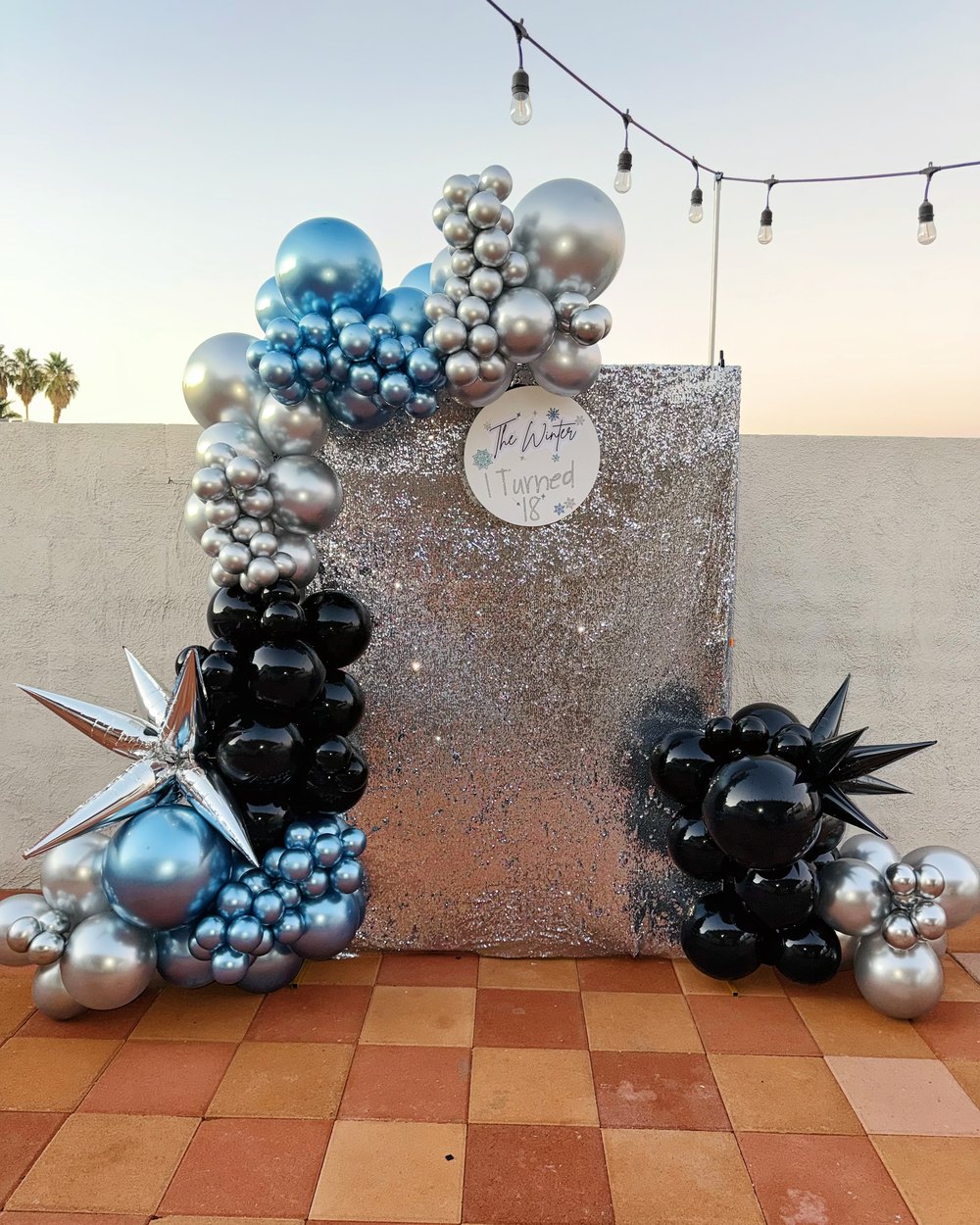 A sequin square backdrop with silver, blue and black balloons and star bursts at sunset