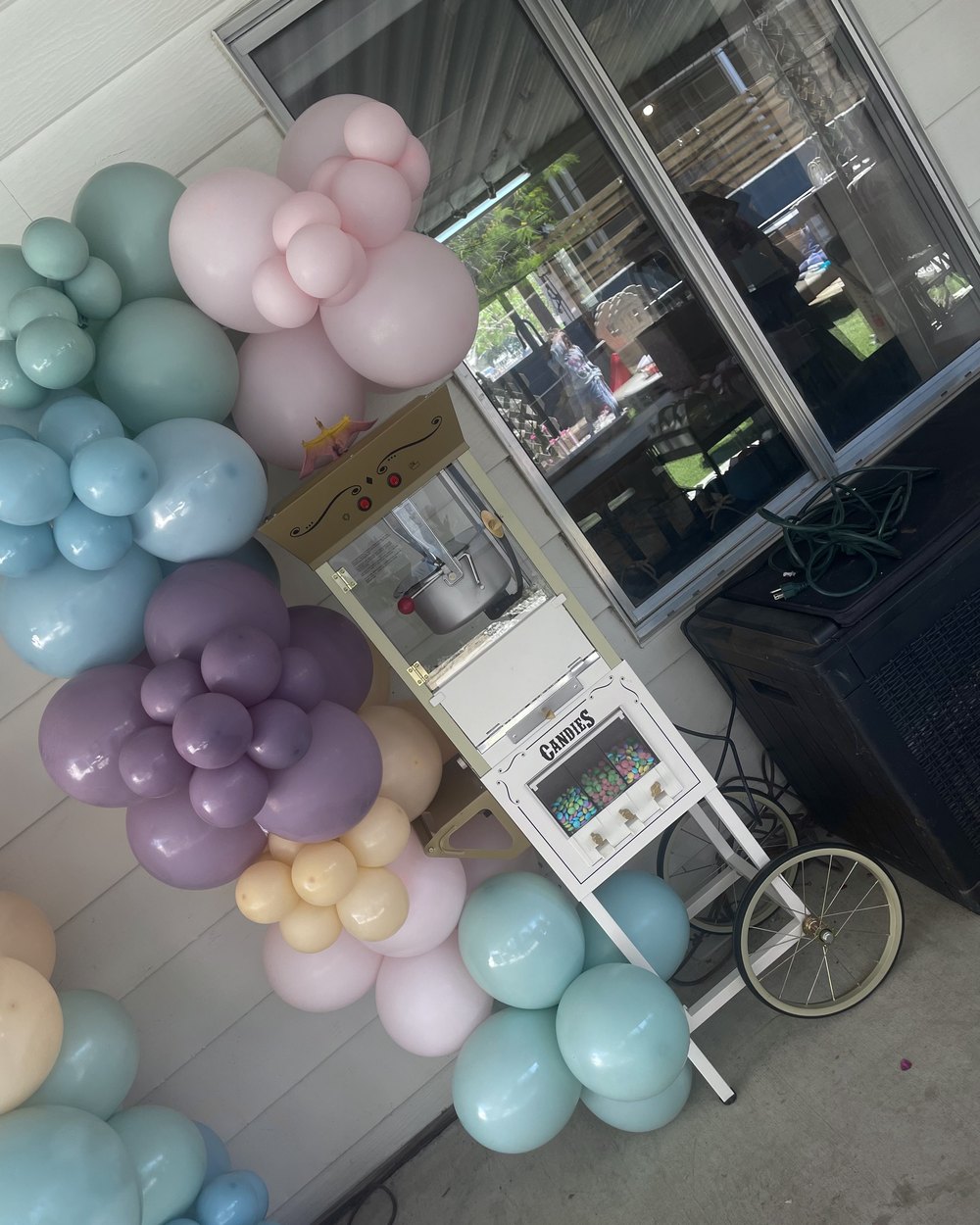 A popcorn machine on a white cart with a candy dispenser, dressed with a pastel balloon garland